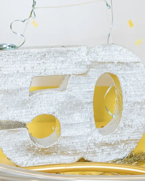 Primer plano de la piñata plateada con forma del número 50, de flecos metalizados, con hilos plateados y globos alargados alrededor.