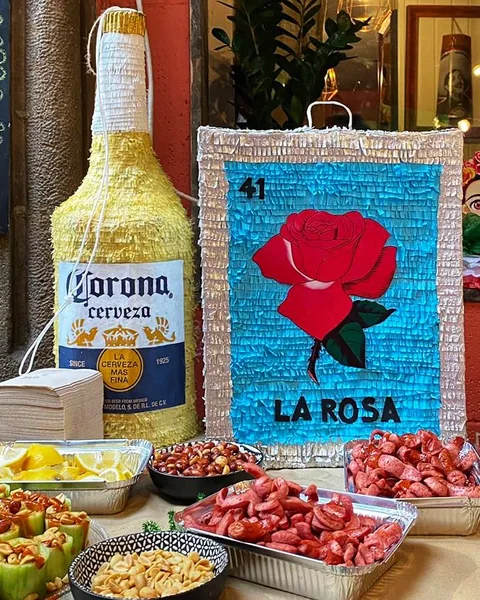 Piñata de una botella de Corona y piñata de la carta de lotería «La Rosa» (número 41, una rosa roja sobre fondo turquesa), sobre una mesa de fiesta con cacahuetes, pepino con chile y limón.