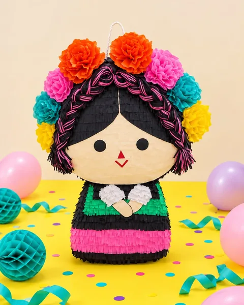 Piñata de muñeca mexicana con trenzas negras y rosas, corona de flores de papel naranjas, rosas, turquesas y amarillas y vestido de rayas verdes, negras y rosas, sobre una mesa amarilla con globos, farolillos turquesa y confeti.