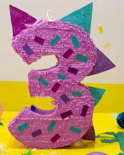 Piñata rosa con forma del número 3, cubierta de confeti de papel morado y turquesa y con picos de colores por detrás como el lomo de un dinosaurio, sobre una mesa amarilla con globos rosas y cintas.